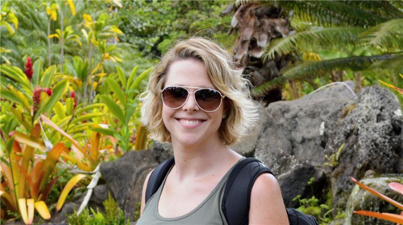 Smiling woman with short blonde hair wearing sunglasses, standing in a lush tropical garden with vibrant plants and rocks, representing team member Maggie Cartie from Oro Valley Dental Arts.